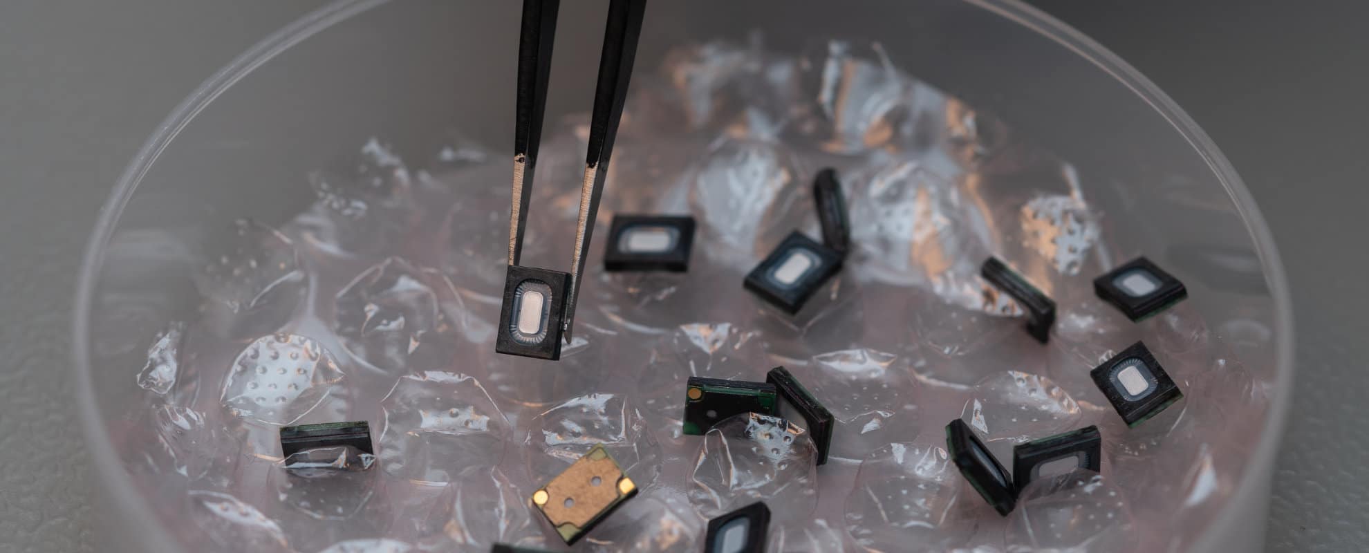 A close-up photograph showing several small USound Ganymede MEMS speakers scattered in a container filled with protective packaging material. In the center, a pair of precision tweezers holds one of the tiny rectangular components, highlighting its compact size and detailed structure. The components have a metallic finish with visible circuitry and openings, reflecting advanced microengineering. The soft lighting and shallow depth of field emphasize the precision and sophistication of the technology. The image conveys innovation, high-quality manufacturing, and the cutting-edge design of USound’s Ganymede MEMS speakers.