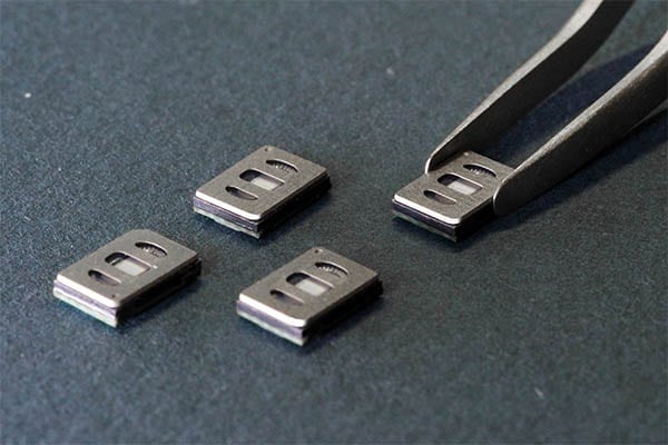 A close-up image showing four small rectangular USound MEMS speakers placed on a dark surface. A pair of precision tweezers is holding one of the components, highlighting its compact design and fine engineering details. Each speaker features a sleek metallic casing with small openings that reveal part of its internal structure. The lighting emphasizes the smooth texture and precision manufacturing of the components. The image conveys innovation, micro-scale technology, and the advanced craftsmanship behind USound’s MEMS speaker products.