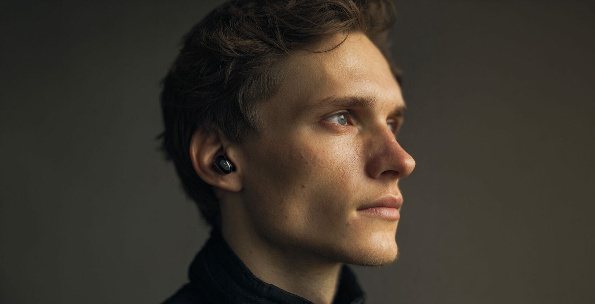 Man wearing wireless in-ear headphones, listening attentively while looking to the side in a quiet indoor environment.
