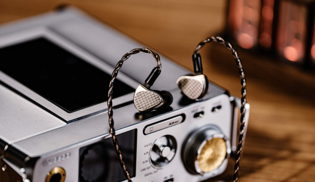 Close-up of Kiwi Ears Halcyon in-ear monitors with braided cable resting on a desktop audio device, highlighting the metallic faceplate and premium build.